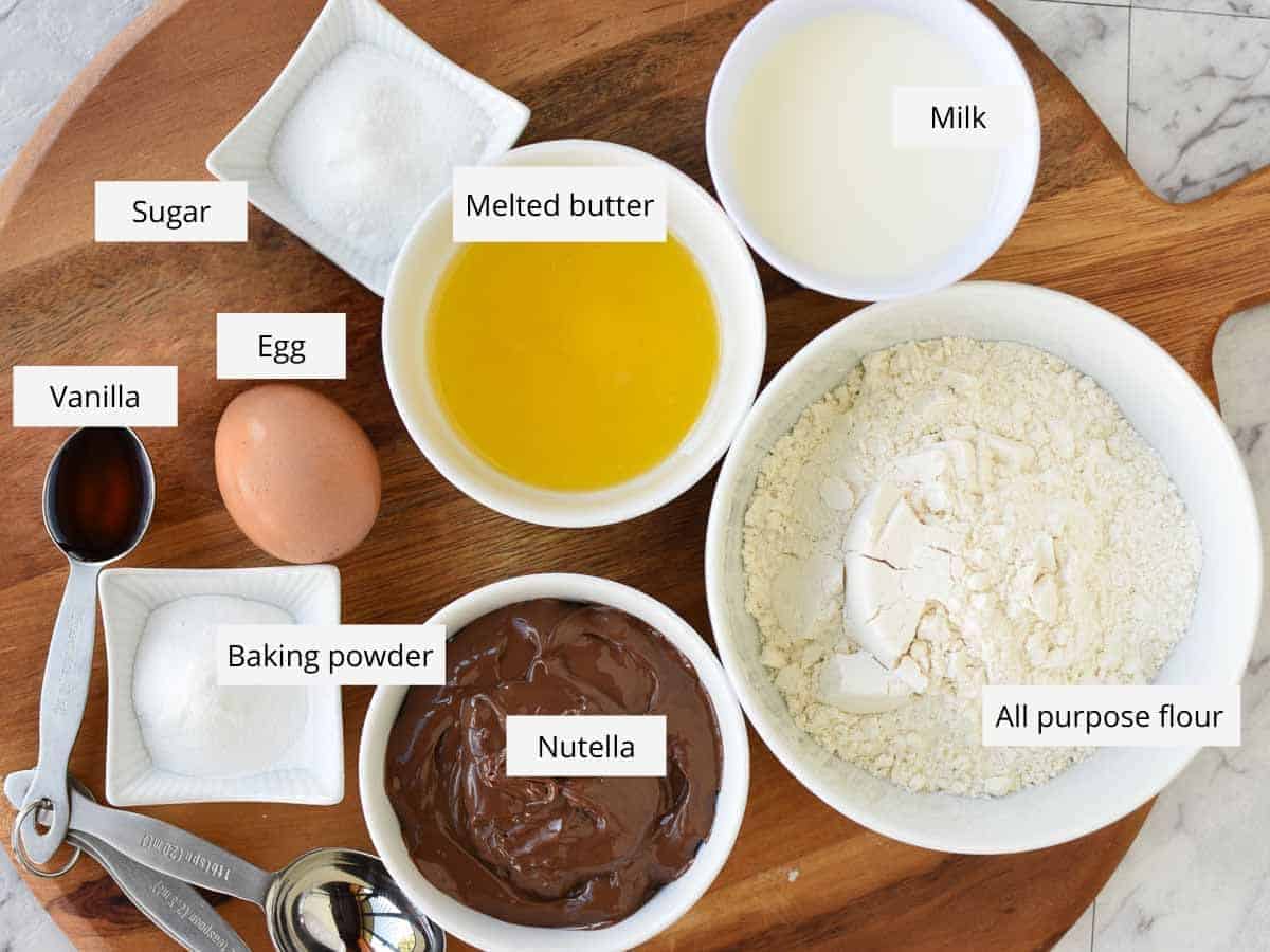 Sugar, melted butter, milk , flour, nutella, baking powder, egg and vanilla in bowls viewed from above.