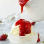 glass bowl of vanilla ice cream with strawberry sauce poured over from a white jug