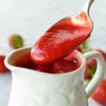 a spoon full of strawberry sauce being poured into a white jug full of more strawberry sauce