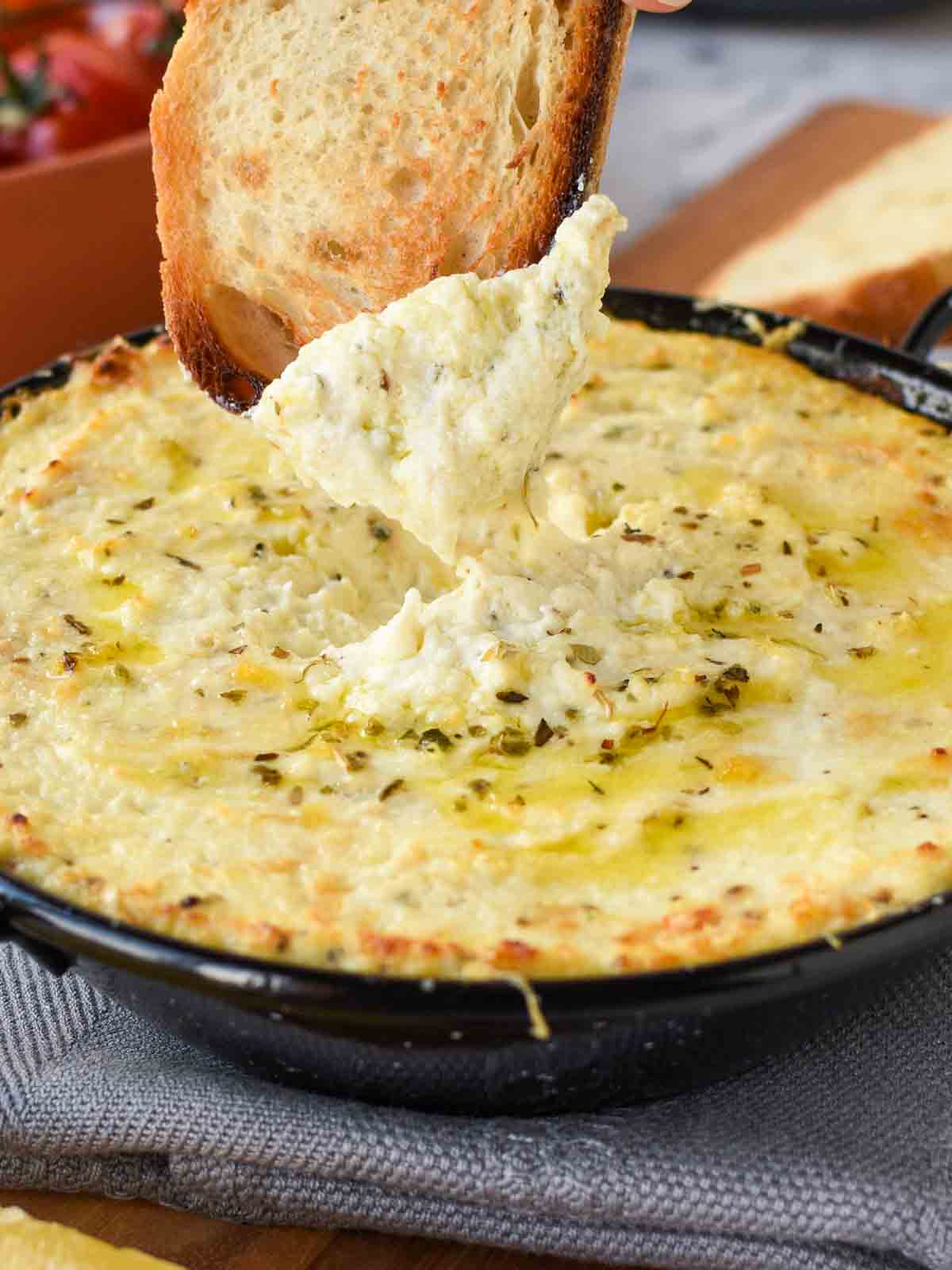 baked ricotta in black enamel pan on grey cloth with a slice of toasted bread dipping in.