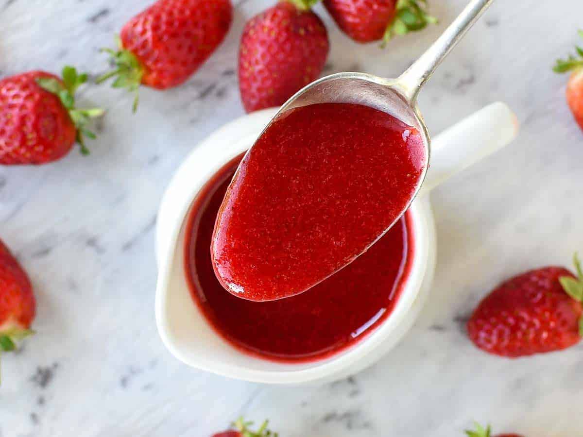 a spoon full of strawberry sauce being poured into a white jug viewed from above.