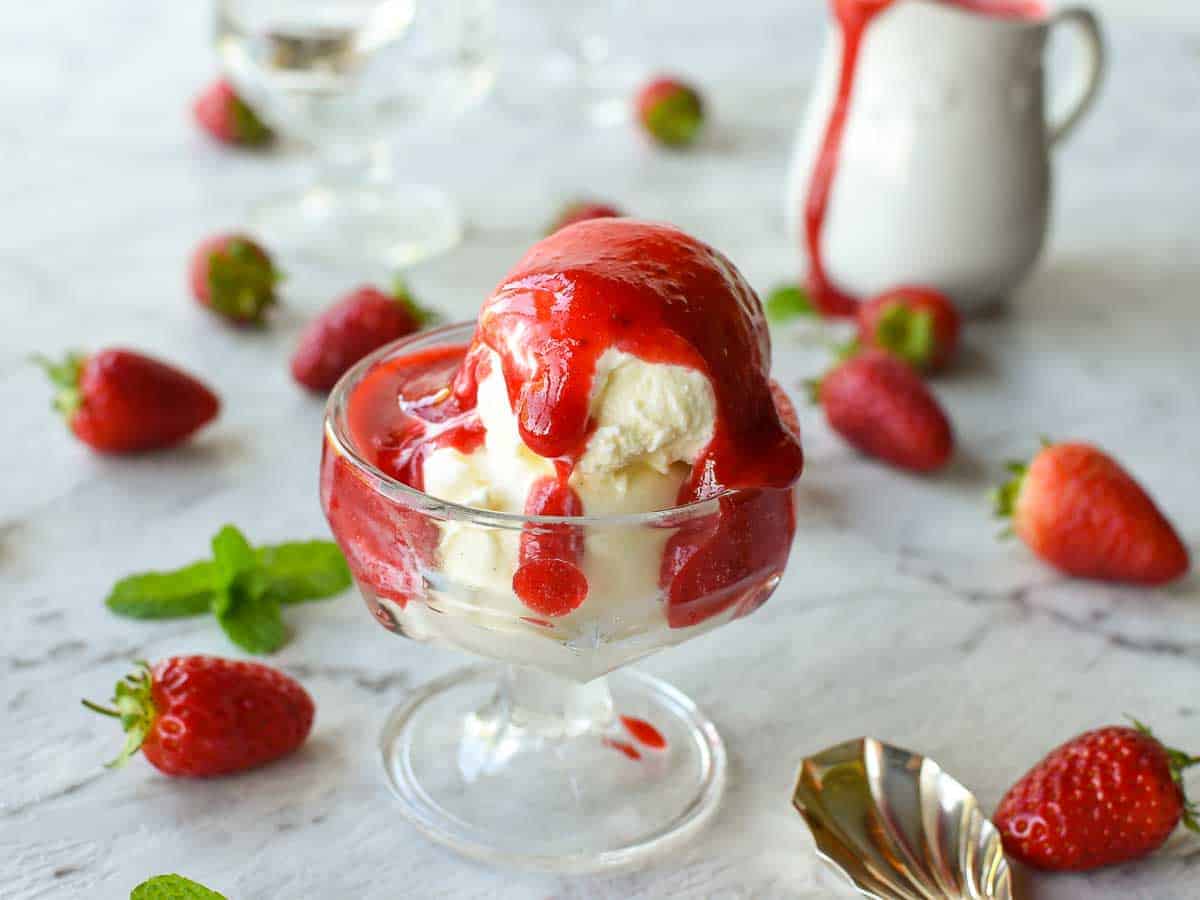 glass bowl of vanilla ice cream with strawberry coulis on top; white jug in the background with a dribble of coulis on the jug.