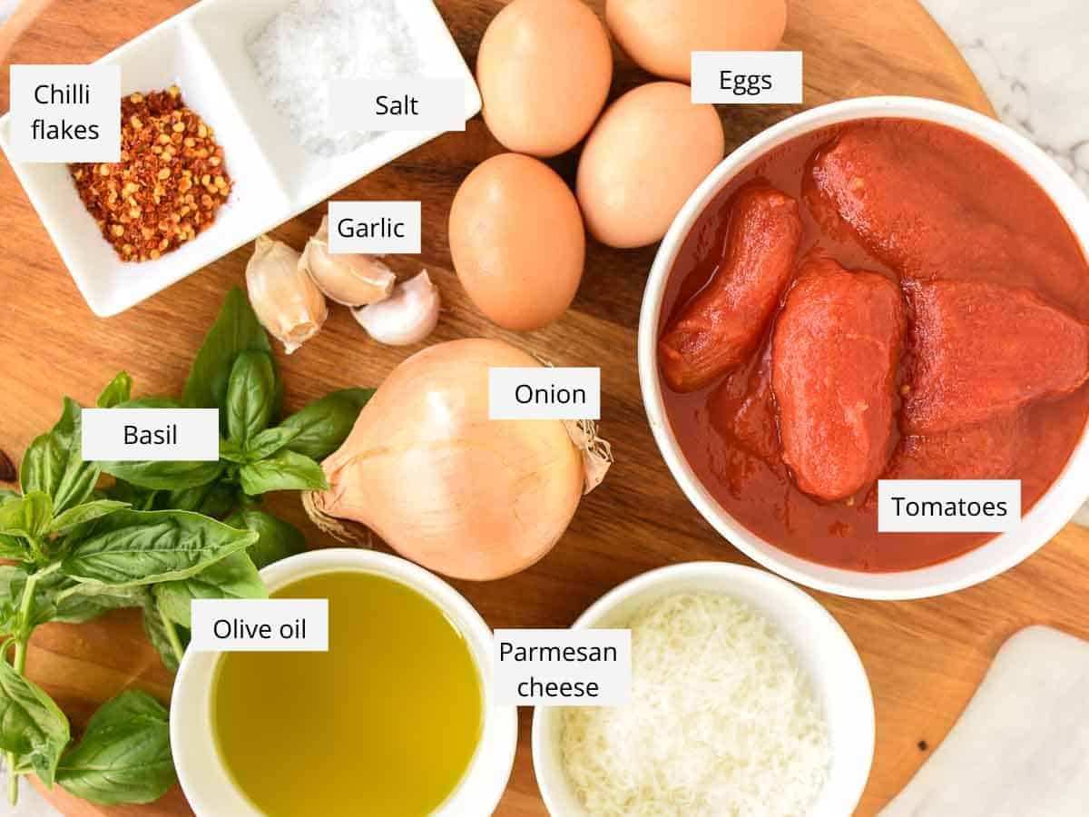 chilli flakes; salt; eggs; tinned tomatoes; grated parmesan cheese; oil; fresh basil; garlic on wooden board viewed from above.