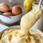 Image with text. Text reads Italian Pastry Cream Thick and Creamy. Image is white bowl of pastry cream with a spoonful being poured in on pale blue cloth with bottle of milk, a lemon, eggs and a vanilla bean in the background