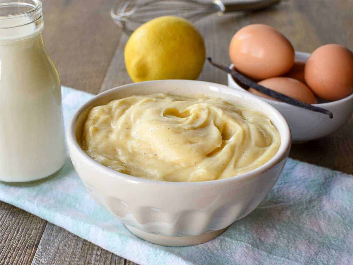 white bowl of pastry cream on pale blue cloth with bottle of milk, a lemon, eggs and a vanilla bean in the background.