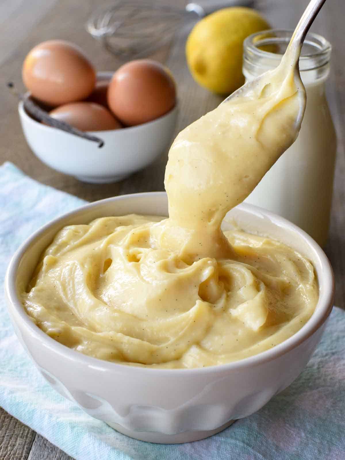 white bowl of pastry cream with a spoonful being poured in on pale blue cloth with bottle of milk, a lemon, eggs and a vanilla bean in the background.