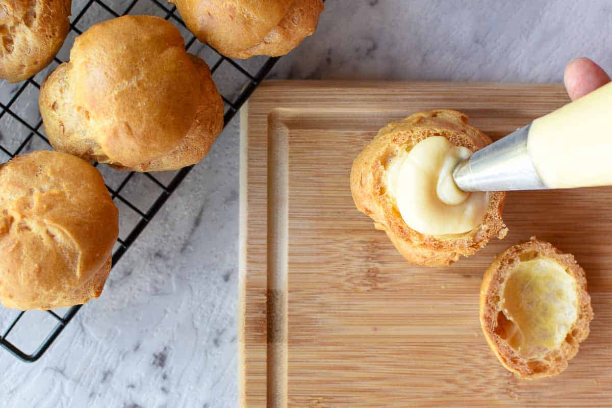 choux pastry being filled using a piping bag on a wooden board; unfilled pastries on black wire rack on the side
