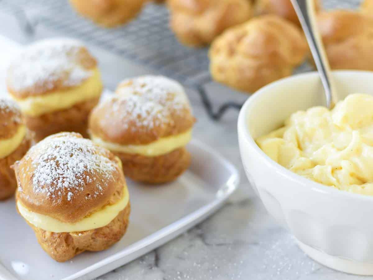 pastries filled with custard on white plate, white bowl of custard and unfilled pastries in the background