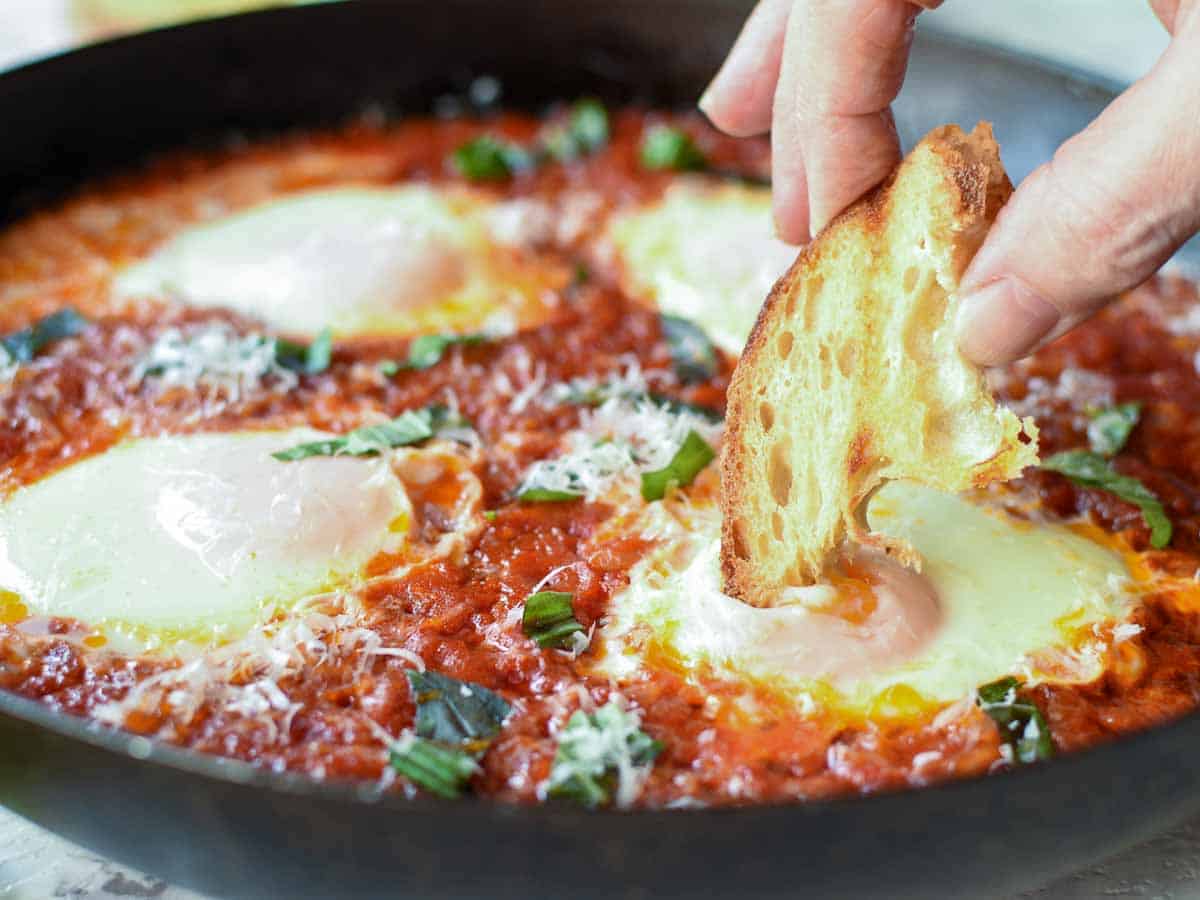 Poached eggs in tomato sauce in black pan, fingers holding toasted bread dipping into egg