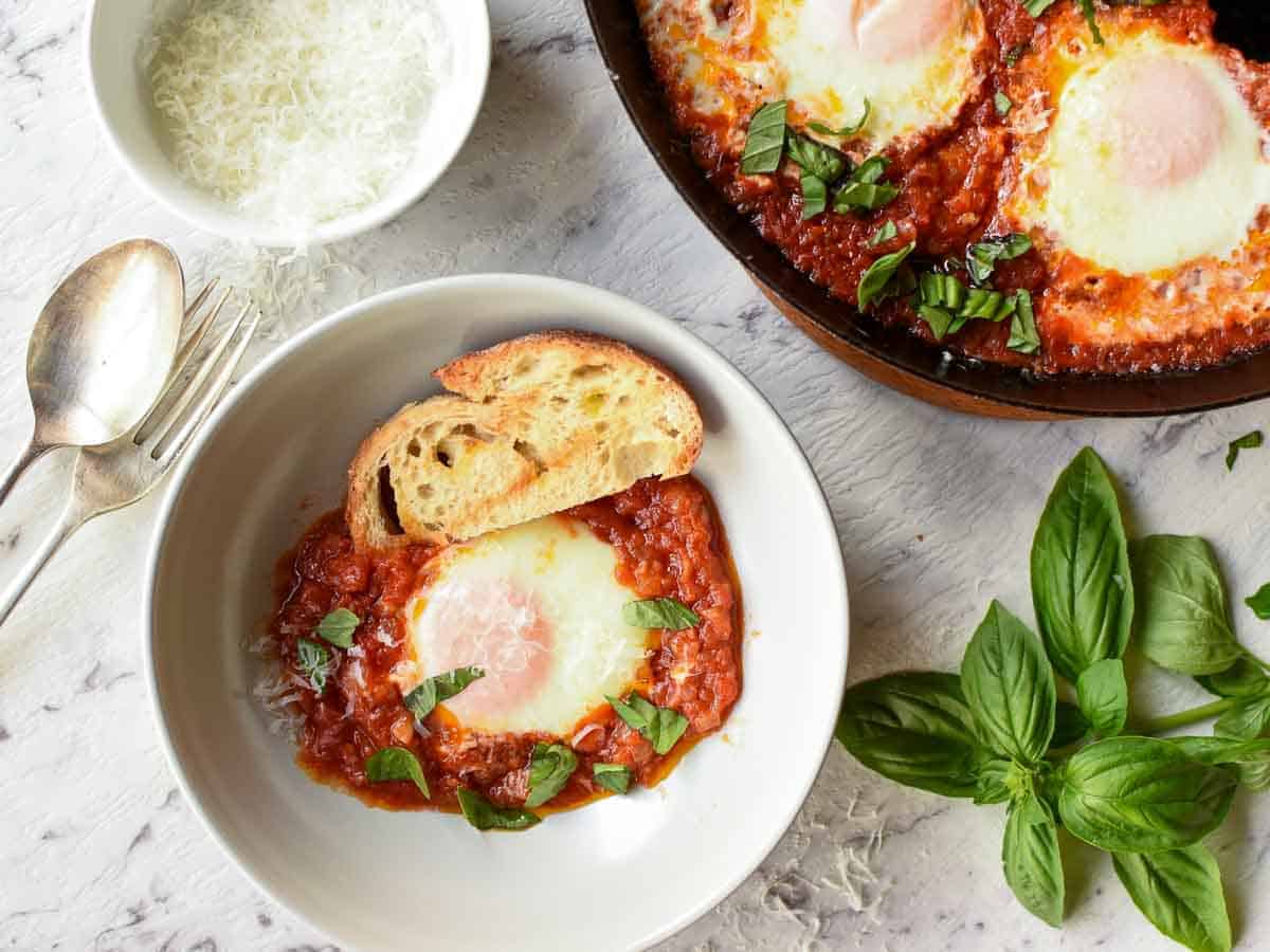 poached egg in tomato sauce and slice of toasted bread in grey bowl, black pan of eggs and tomato sauce partly out of the picture viewed from above.