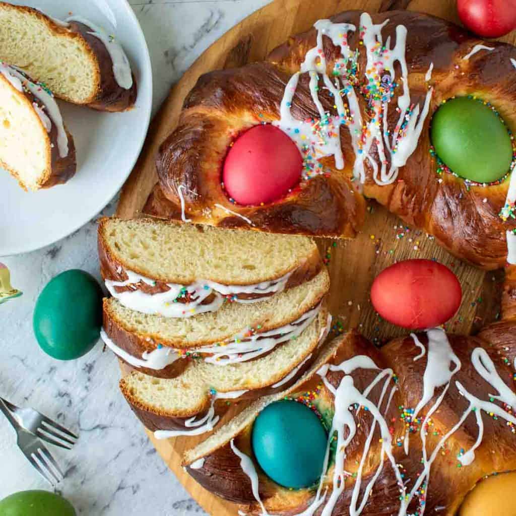 close up of baked sweet bread decorated with red, blue, yellow and green eggs and drizzle of icing and a few slice cut to show inside of bread. In the top left hand corner is two slice of the bread on a white plate. Viewed from above