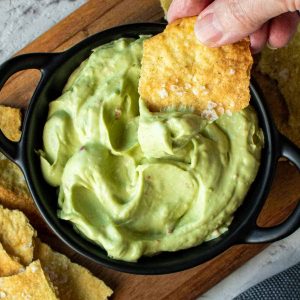 cracker with avocado dip on being scooped out of avocado dip in black bowl viewed from above