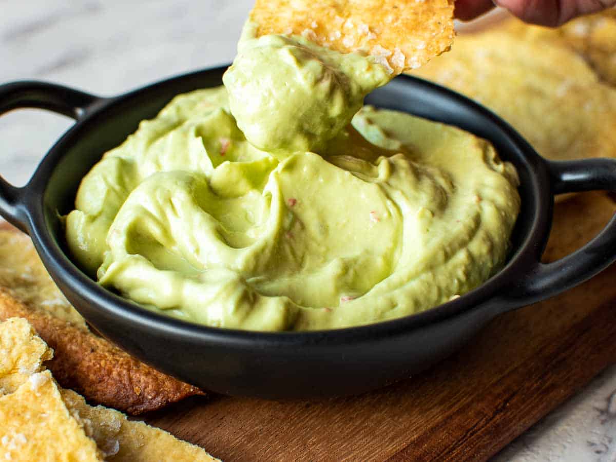 cracker with avocado dip on being scooped out of avocado dip in black bowl.
