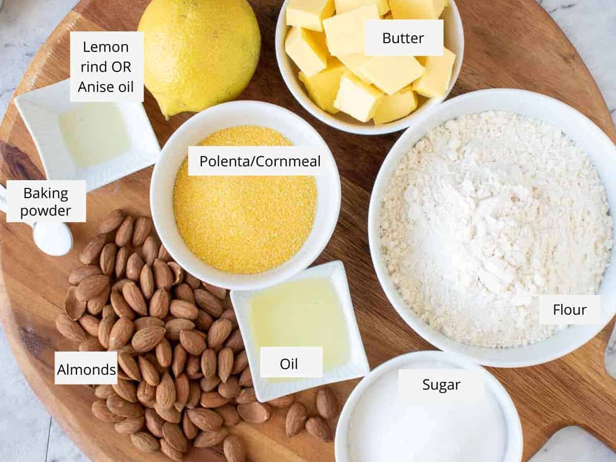 butter, flour, sugar, oil, almonds, baking powder, anise oil, lemon and cornmeal in bowls on wooden board.