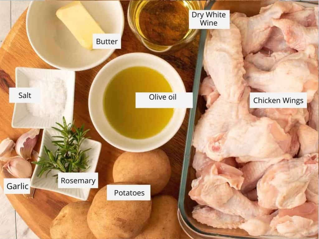 Butter, dry white wine, raw chicken wings, potatoes, rosemary, garlic, salt and olive oil in bowls and dishes on a wooden board