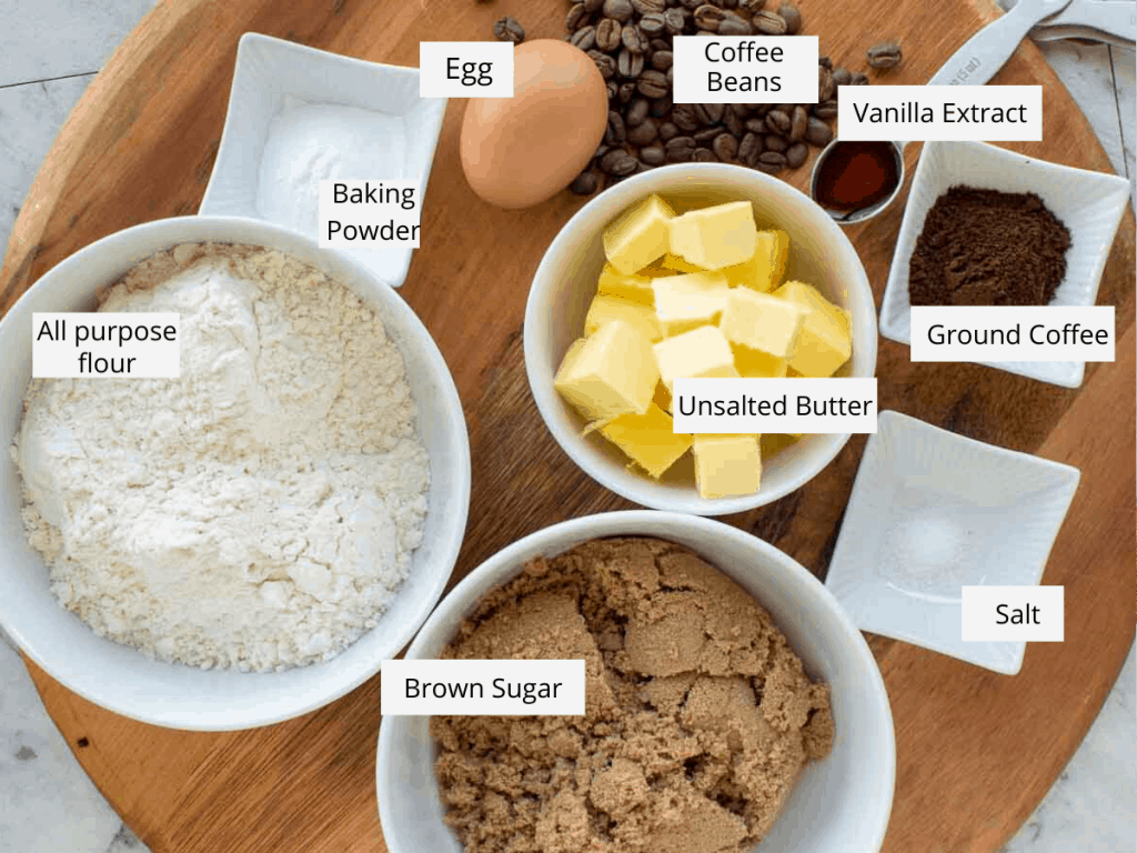 flour, baking powder, an egg, coffee beans, vanilla extract in a spoon, ground coffee, salt, brown sugar and unsalted butter in white bowls all on wooden board