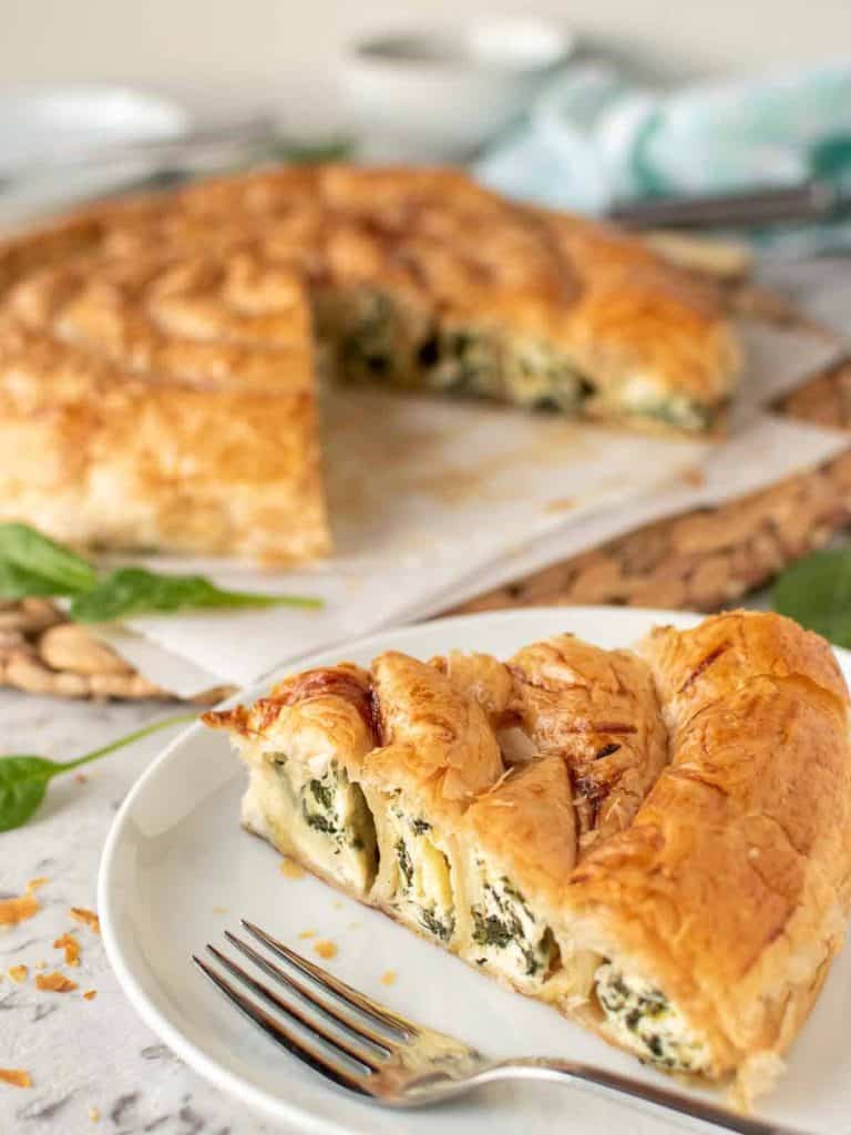 slice of spinach ricotta filo pie on white plate with remaining pie in the background