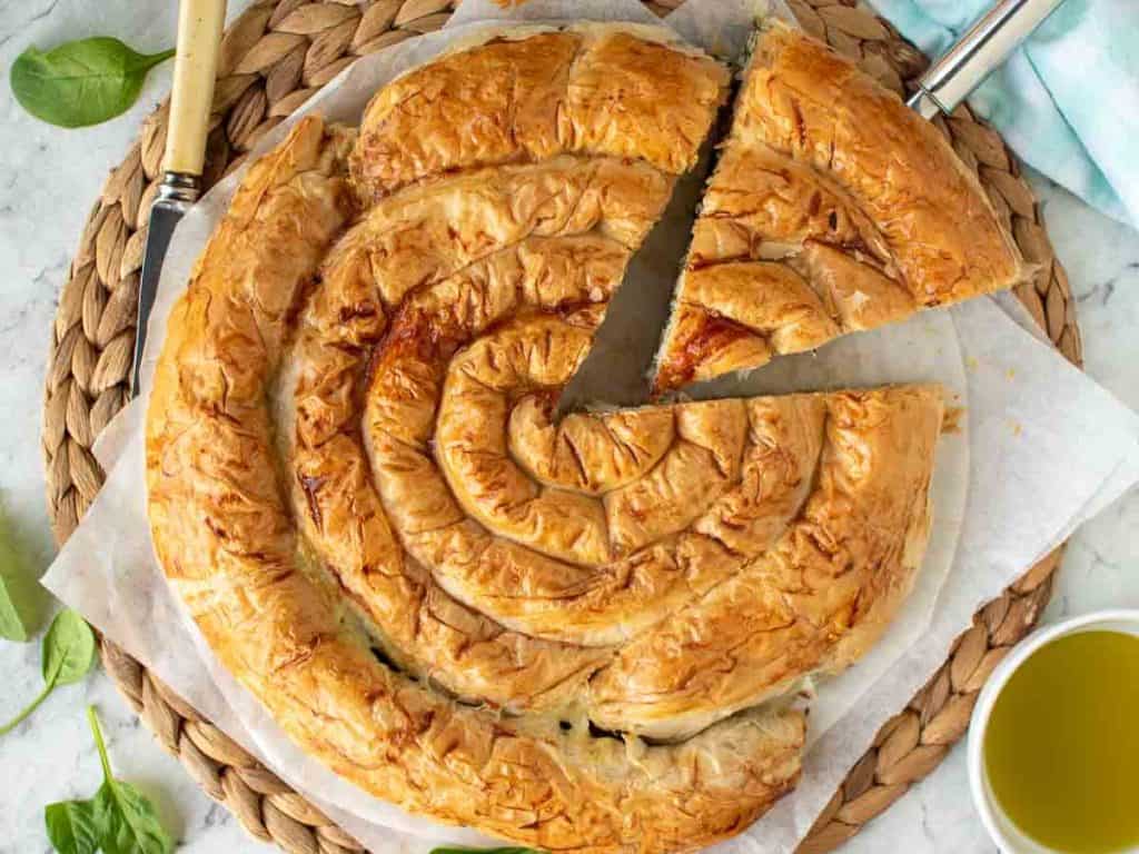 spiral filo pie with a slice cut on plaited raffia mat with bowl of oil and spinach leaves scattered around viewed from above