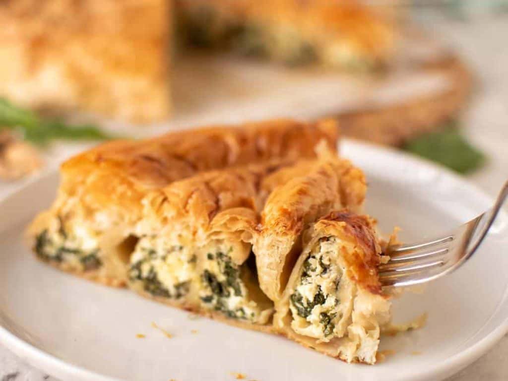 slice of spinach ricotta filo pie on white plate with fork piercing the edge