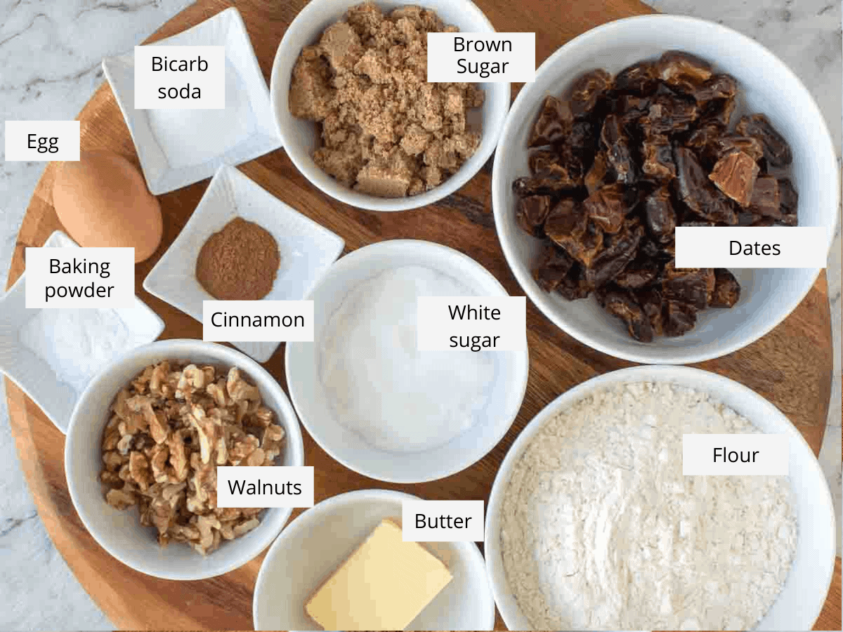 ingredients on wooden board - baking powder, brown and white sugar, dates, flour, butter, walnuts, baking soda, egg and cinnamon
