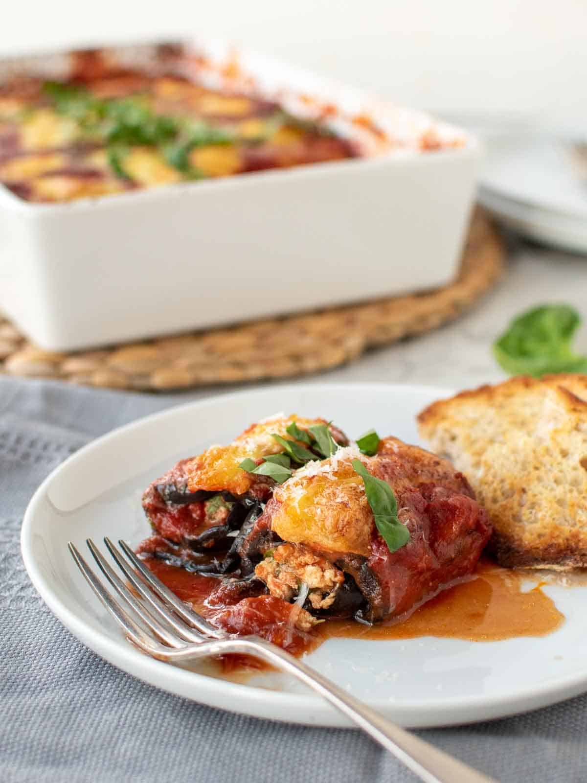 two eggplant involtini on a white plate with fork with white baking dish in the background.