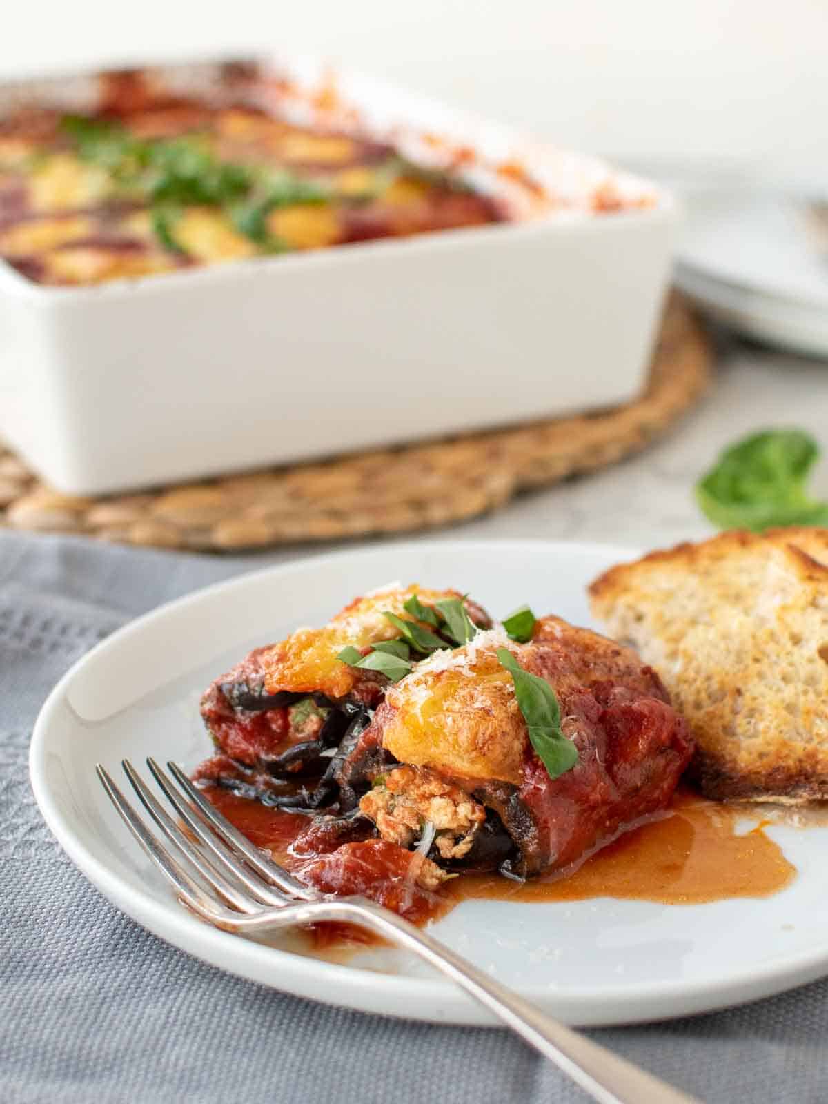 Two eggplant involtini on a white plate with fork with white baking dish in the background.