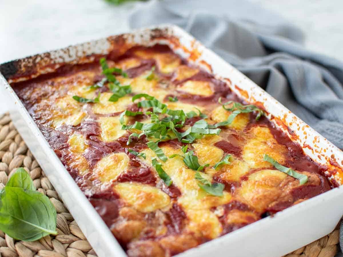 White baking dish filled with tomato sauce and cheese topped with green herbs.