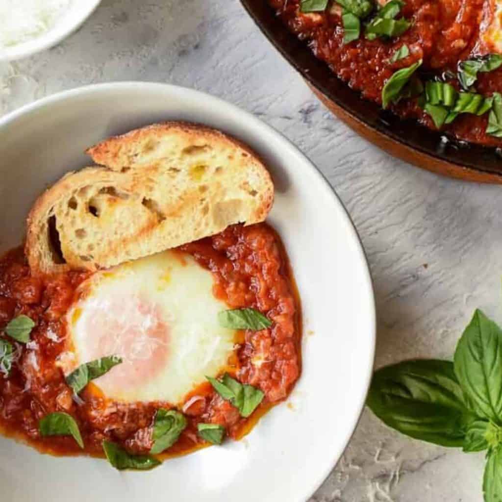 poached egg in tomato sauce and slice of toasted bread in grey bowl, black pan of eggs and tomato sauce partly out of the picture viewed from above