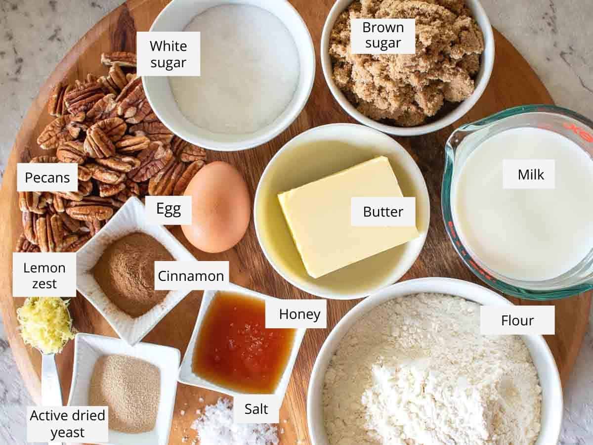 ingredients arranged on wooden board - white and brown sugar, milk, flour, salt, honey, butter, yeast, cinnamon, lemon zest, egg and pecans