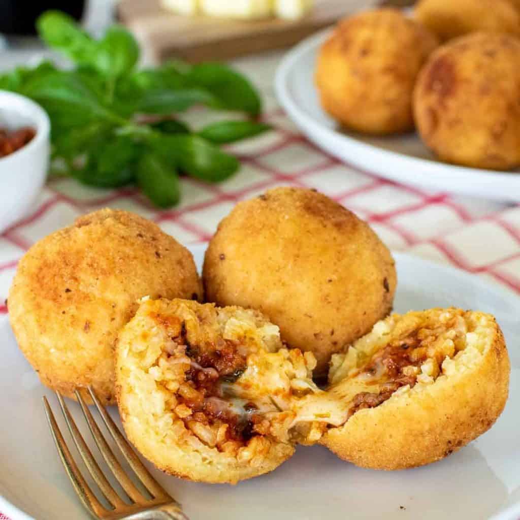 2 whole arancini and one split on a white plate with fork; arancini and basil in the background