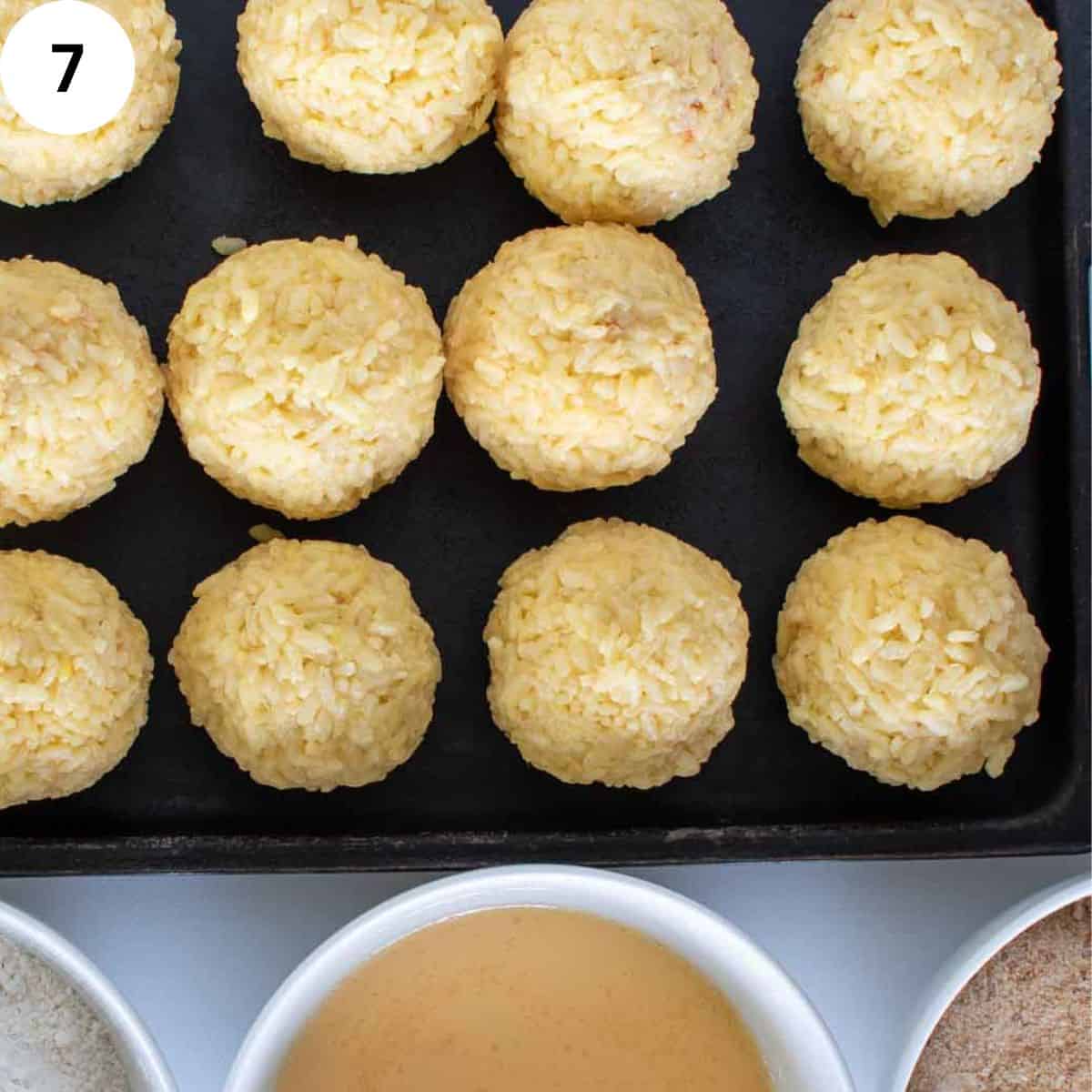 Rice balls lined up on baking sheet with bowls of egg and bread crumbs.