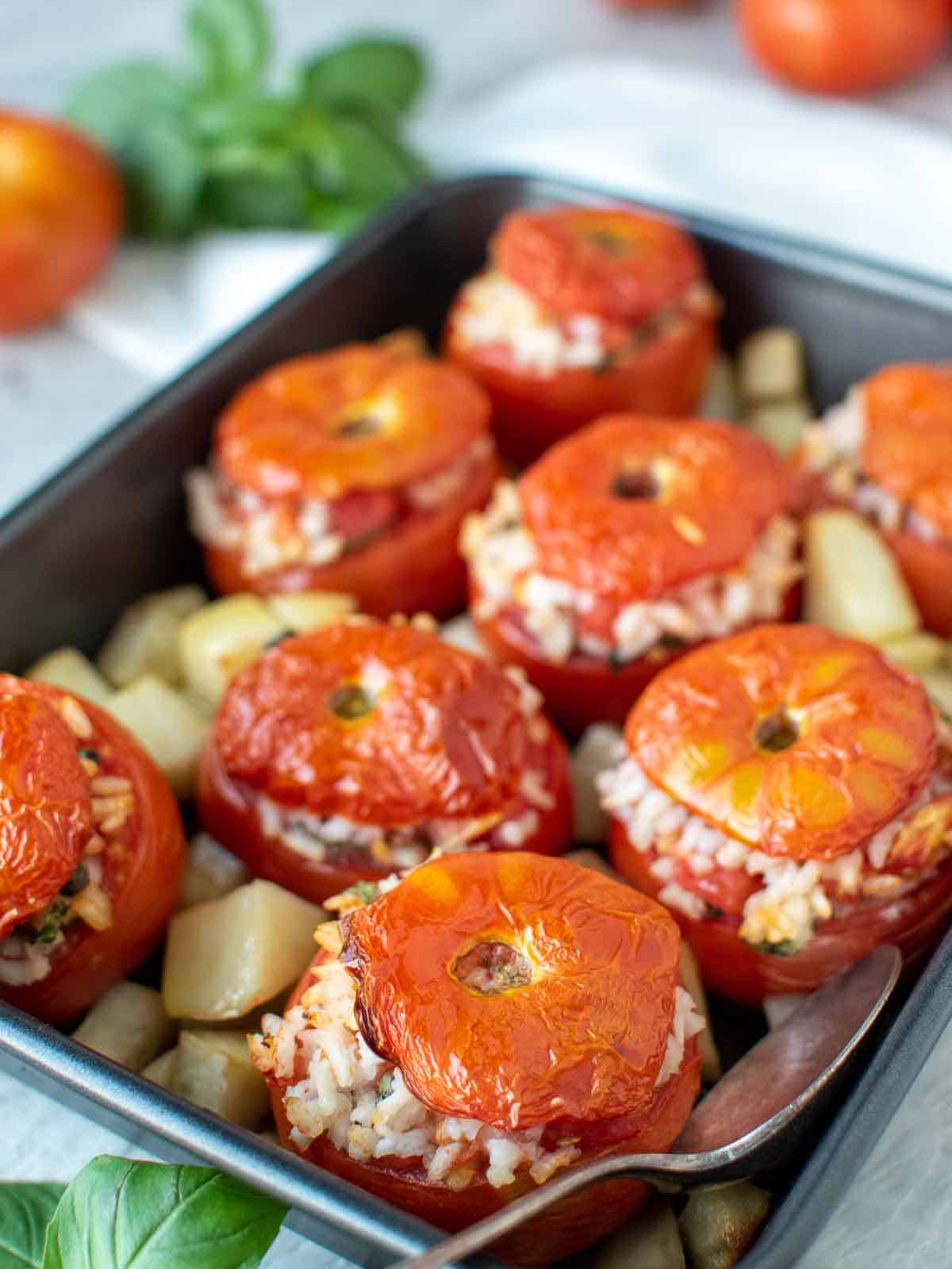 eight baked tomatoes stuffed with rice in black baking pan with cubed roasted potatoes.