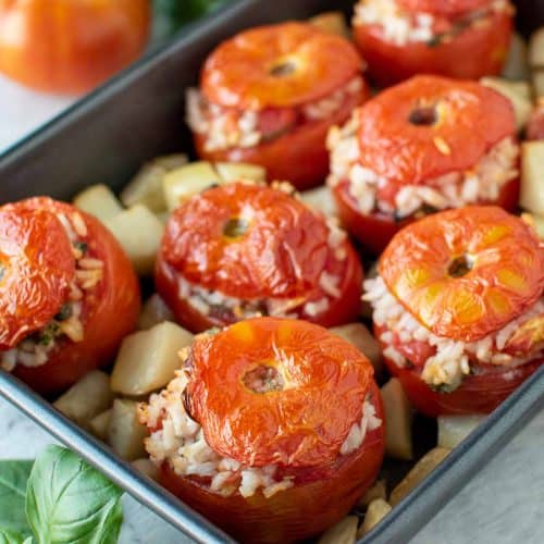eight baked tomatoes stuffed with rice in black baking pan with cubed potatoes.