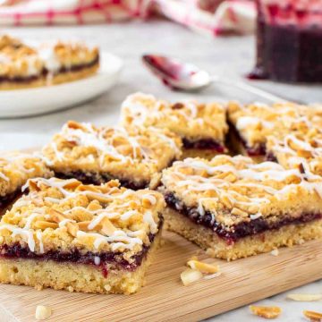 raspberry bars on wooden board with jam and extra bars in the background.