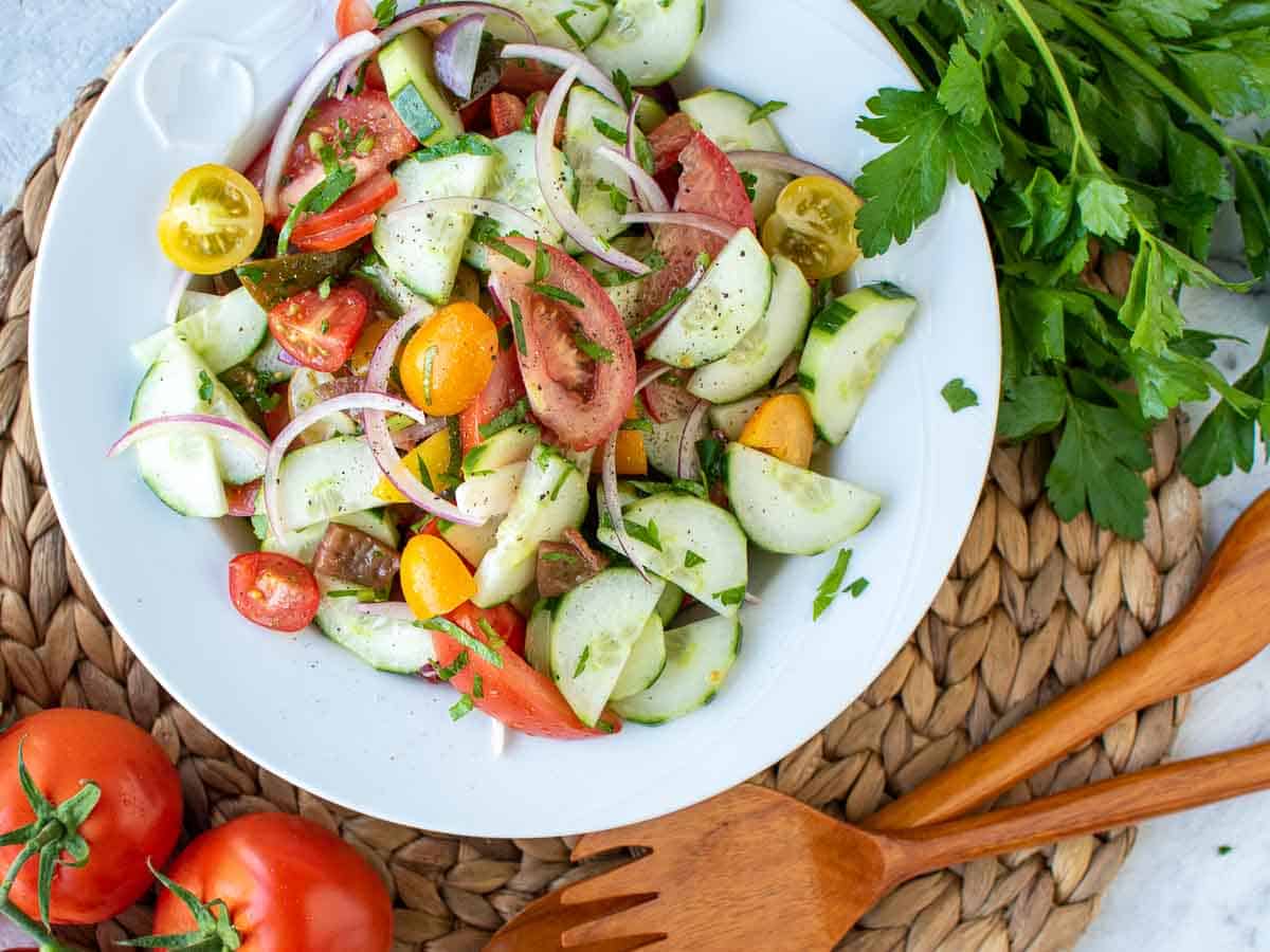 cucumber tomato onion salad on white plate with tomatoes and parsley nearby viewed from above.