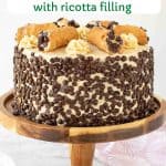 image with text overlay. text reads the best cannoli cake with ricotta filling. image is cake covered in mini choc chips with 6 cannoli on top. cake on on a wooden cake stand.