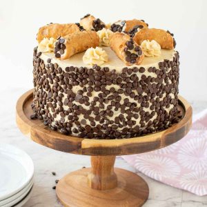 cake covered in mini choc chips with 6 cannoli on top. cake on on a wooden cake stand.