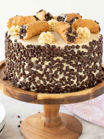 cake covered in mini choc chips with 6 cannoli on top. cake on on a wooden cake stand.