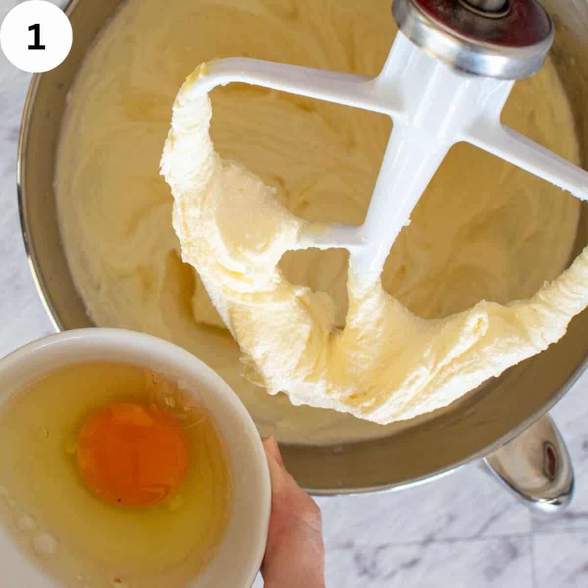 Creamy mixture clinging to mixer paddle and a cracked egg in a bowl.