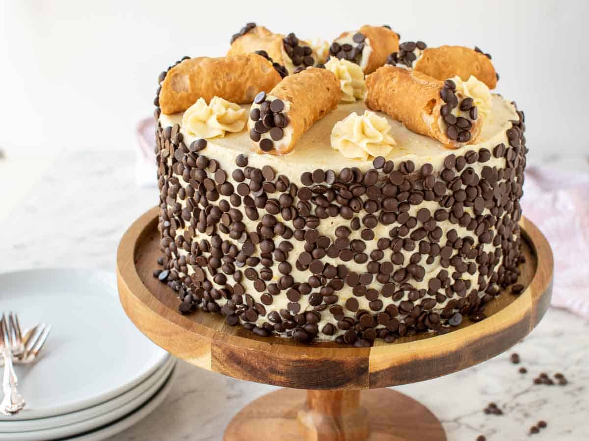 Close up of a cake with sides covered in chocolate chips with six filled cannoli on top.