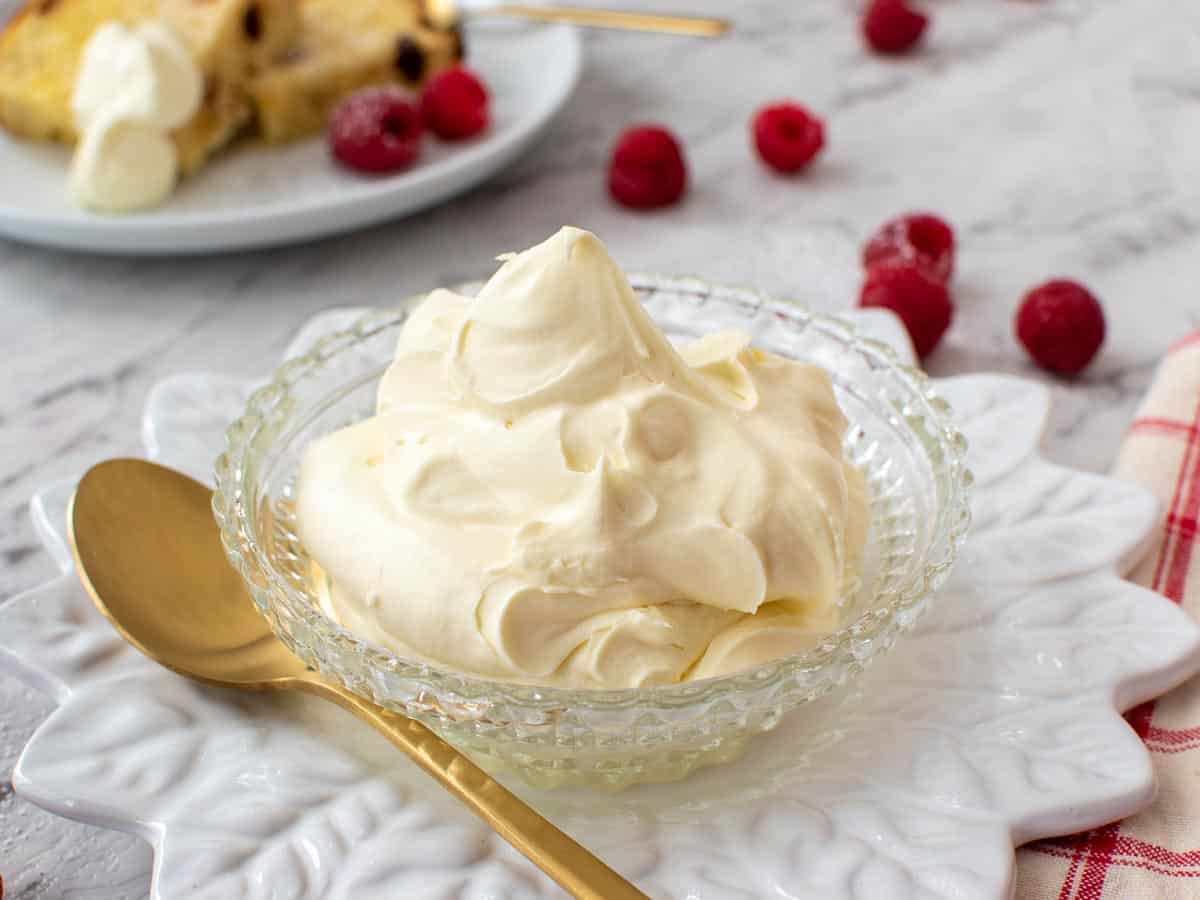cream in a bowl with gold spoon, raspberries scattered around and plate with cake and cream in the background