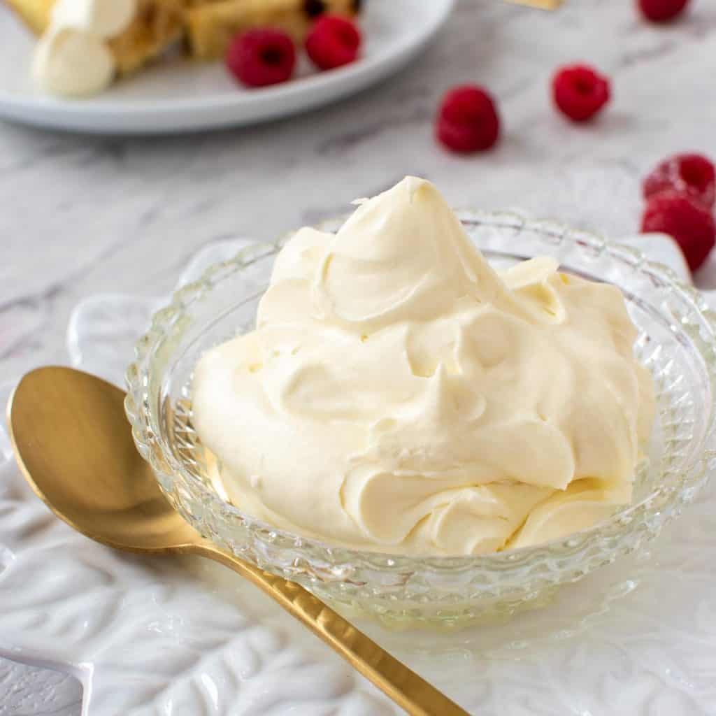 close up of mascarpone cream in a bowl with gold spoon, raspberries scattered around and plate with cake and cream in the background