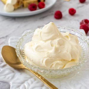 close up of mascarpone cream in a bowl with gold spoon, raspberries scattered around and plate with cake and cream in the background