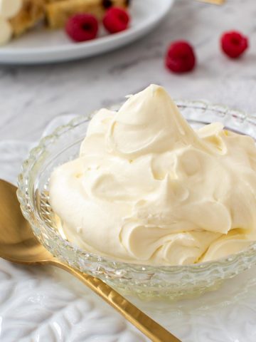 close up of mascarpone cream in a bowl with gold spoon, raspberries scattered around and plate with cake and cream in the background