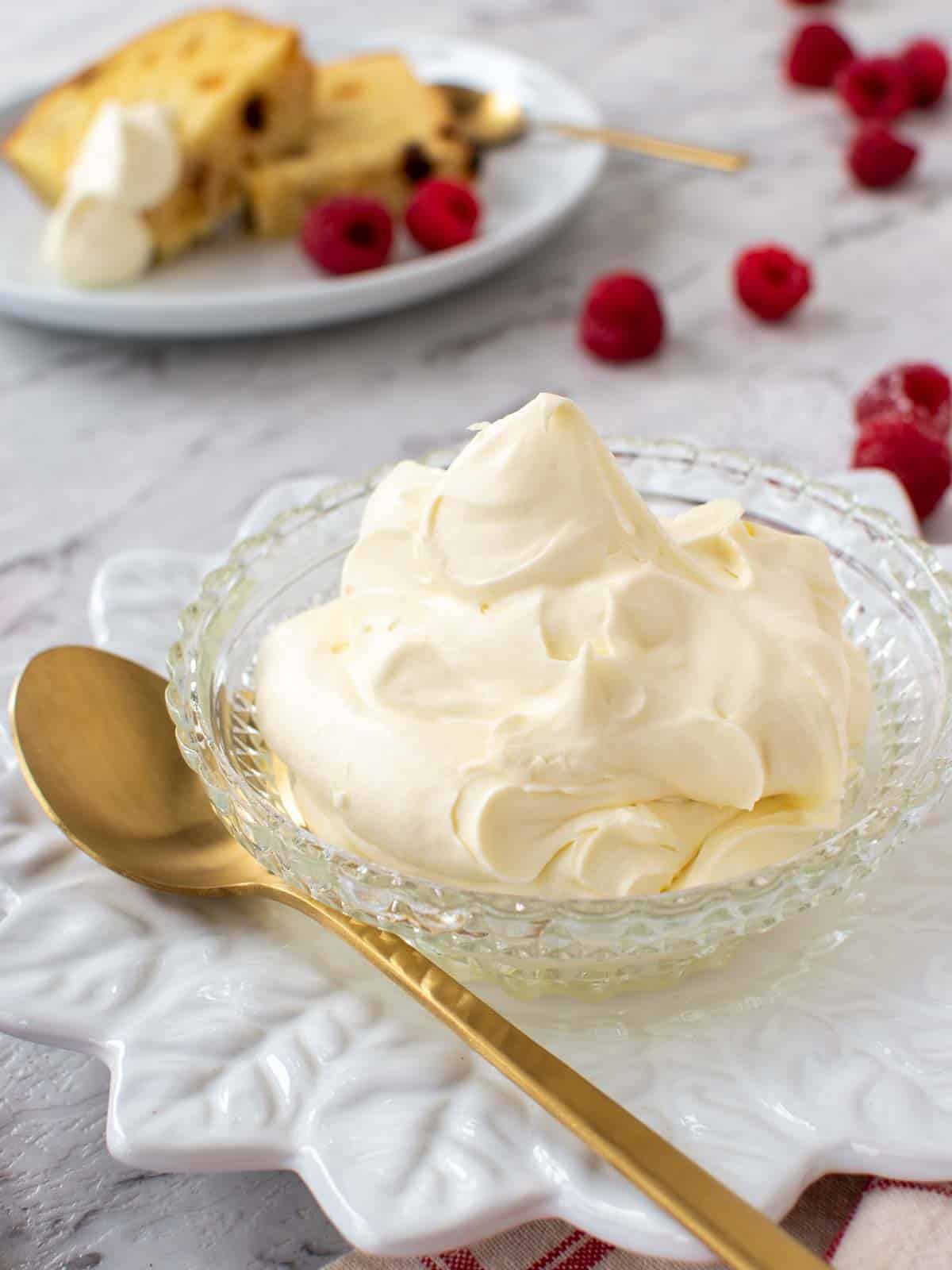 mascarpone cream in a bowl with gold spoon, raspberries scattered around and plate with cake and cream in the background.