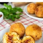 2 whole arancini and one split on a white plate with fork; arancini and basil in the background