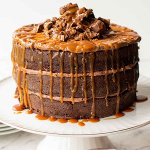 three layer chocolate cake with chocolate frosting and dripping caramel on a white cake stand.