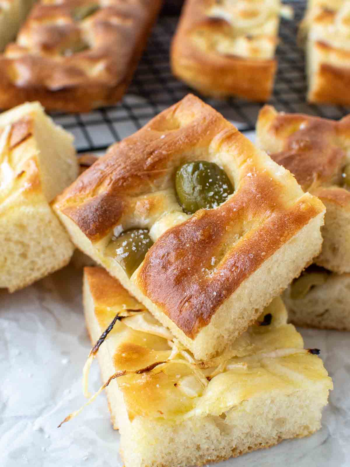 close up of focaccia genovese with green olives on top and sliced onions on other pieces with more focaccia in the background.