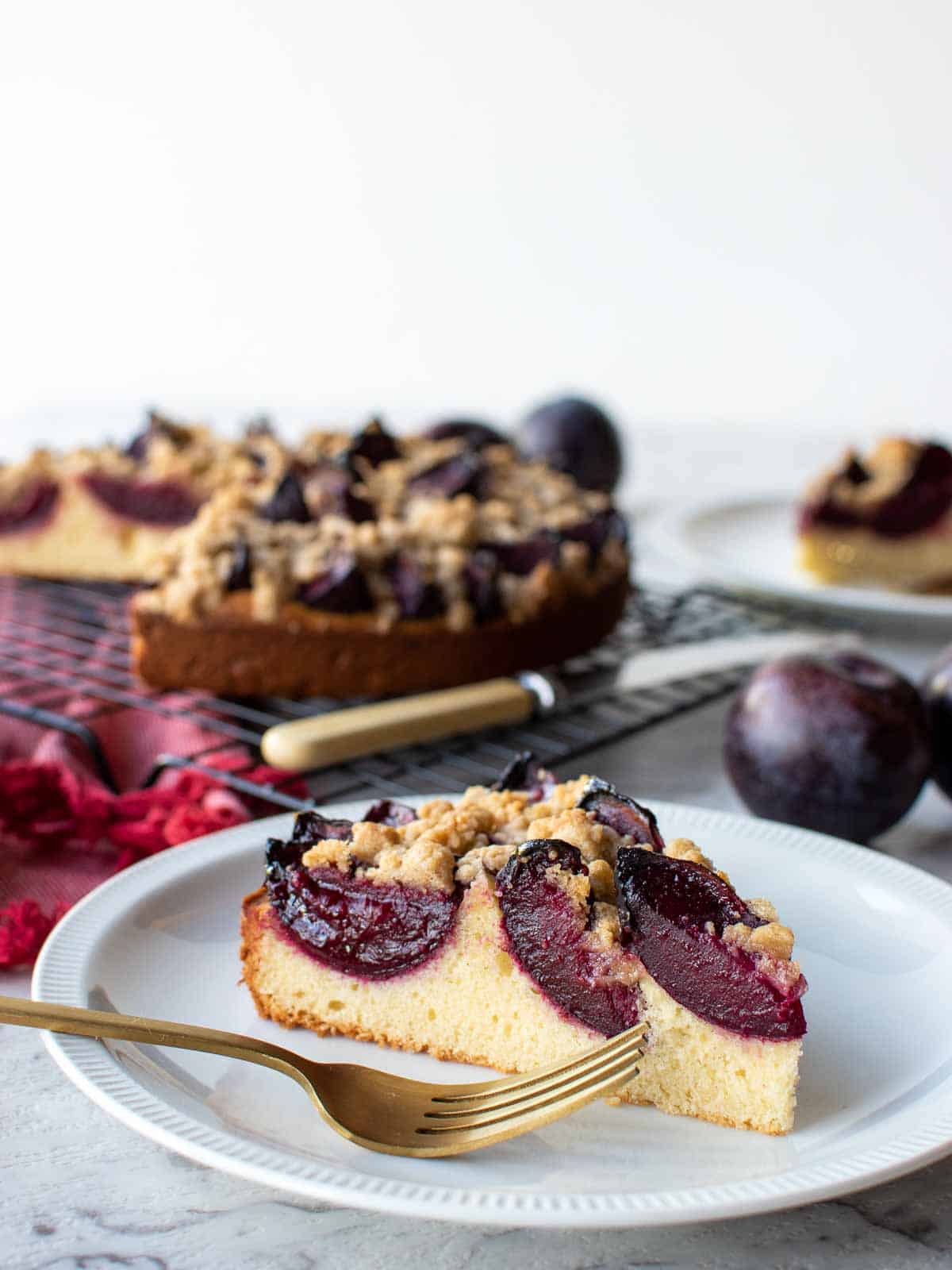 A slice of German plum cake on white plate with whole cake in the background.