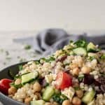 Image with text. text reads "how to make greek couscous salad. image is bowl of couscous salad with fork and spoon in the front and grey towel in the background.